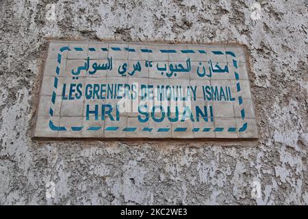 Sign outside the Heri es-Souani royal granary in the city of Meknes, Morocco. Heri es Souani (Heri es-Souani) is an ancient Royal granary built in the 17th Century by Sultan Moulay Ismail. The royal granary was built to withstand a decade-long siege and had massive walls (4m thick) with arched ceilings and an underground fresh water system. The high ceilings and small windows kept the complex cool in the summer and warm in the winter, and helped air circulation (important for preventing grain spoilage). The granary stored grain and hay for the adjacent royal stables. An army of cats helped wit Stock Photo