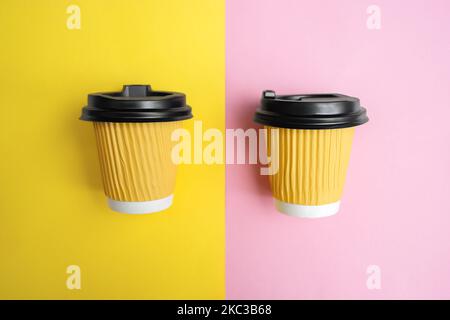Two yellow cups for takeaway coffee or tea lie parallel on colored paper on a design table. Orange and pink background. Copy space for text. Soft Selective focus Stock Photo