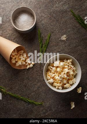 White porcelain bowl and brown paper cone full of popcorn with salt and green sprigs of rosemary on marble countertop Stock Photo