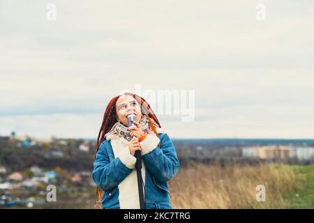 Young woman with dreadlocks singing into microphone in countryside. Female singer enjoying singing in nature Stock Photo
