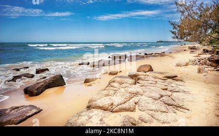 Ebb tide coming in on a rocky seashore. Surf waves break over shallow reef. Cloudy blue sky on a spring day on Sunshine Coast, Queensland. Stock Photo