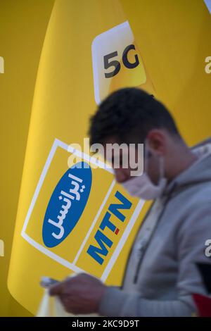 An Iranian man uses his smartphone as he walks past a 5G and MTN-Irancell logos in the sixth Iran’s Internet of Things exhibition (IoTex 2021) in Tehran International Exhibition Center on February 16, 2021. Iran’s first 5G network has ran by the Irancell company which is an Iranian telecommunications company that operates Iran's largest 2G-3G-4G-4.5G also 5G mobile network, and fixed wireless TD-LTE internet services. (Photo by Morteza Nikoubazl/NurPhoto) Stock Photo