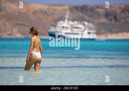 A young woman wearing a white bikini as seen in the sea of Balos with a ferry in the background. Panoramic view of Balos Beach, the incredible lagoon with the turquoise exotic and tropical water of the Mediterranean sea is located in Chania region in Crete Island. Balos is one of the most visited beaches in Crete and popular for visitors around the world. Crystal clear water, the lagoon, rocky steep mountains, a beach bar providing umbrellas and shadow with beverages and a pirate island are located at the same region that is accessible by a 20 min trek or boat. Greece is trying to boost its to Stock Photo