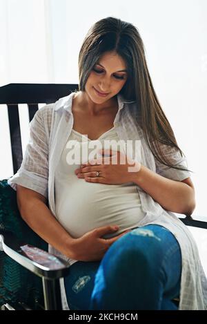 Soon youll be in my arms. a happy pregnant woman cradling her belly while relaxing in a chair at home. Stock Photo