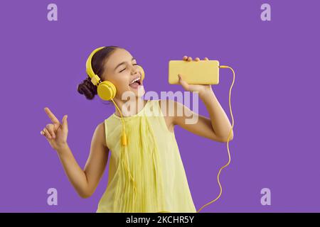 Cheerful preteen girl in headphones connected to mobile phone listens to music and sings along. Stock Photo