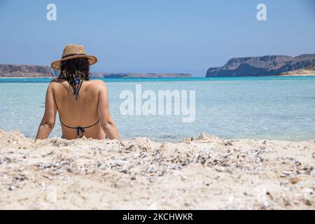 A young woman wearing a hat is sitting on the white pink sand of Balos Beach, the incredible lagoon, with the pool like turquoise exotic and tropical water of the Mediterranean sea is located in Chania region in Crete Island. Balos is one of the most visited beaches in Crete and popular for visitors around the world. Crystal clear water, the lagoon, rocky steep mountains, a beach bar providing umbrellas and shadow with beverages and a pirate island are located at the same region that is accessible by a 20 min trek or boat. Greece is trying to boost its tourism and give privileges to vaccinated Stock Photo