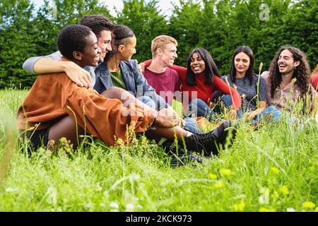 Group of multiethnic friends having fun together in the natural park - young men and women sitting outdoors in the grass hugging and conversing togeth Stock Photo