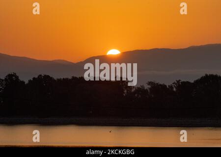 The sun is appearing behind the mountains and is reflecting on the water surface. Magic hour summer sunrise with warm colors of the sky and the sun over Kerkini with birds, horses, local fishermen fishing spotted as silhouettes, at Lake National Park in Serres region in Northern Greece. Artificial Lake Kerkini is a unique wetland, a National Park and protected by the Ramsar Convention as a wetland with thousands of birds including rare and protected, riverside forest as important hydrobiospheres are developing which are of great international significance and acceptance, the main water source  Stock Photo