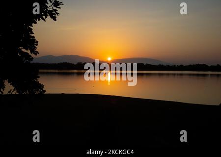 The sun is appearing behind the mountains and is reflecting on the water surface. Magic hour summer sunrise with warm colors of the sky and the sun over Kerkini with birds, horses, local fishermen fishing spotted as silhouettes, at Lake National Park in Serres region in Northern Greece. Artificial Lake Kerkini is a unique wetland, a National Park and protected by the Ramsar Convention as a wetland with thousands of birds including rare and protected, riverside forest as important hydrobiospheres are developing which are of great international significance and acceptance, the main water source  Stock Photo