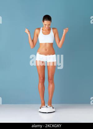 Start strong, finish stronger. Studio shot of a fit young woman weighing herself on a scale and cheering against a blue background. Stock Photo