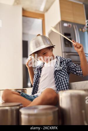 All he needs is a little imagination. a happy little boy playing drums with pots on the kitchen floor while wearing a bowl on his head. Stock Photo