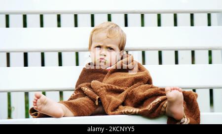 a little one-year-old girl, wrapped in a towel, with a dirty face, lonely sitting on a swing in the garden, in the summer. She has a sad look. She wants to sleep. High quality photo Stock Photo