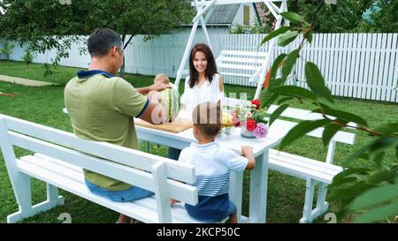 A young family with two little funny children sitting at a dinner table in the garden, in summer. Dad cuts a large watermelon, is going to treat his family. lunch with the family. High quality photo Stock Photo