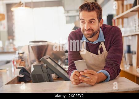 It feels good to be my own boss. a happy business owner using his smartphone while leaning on the counter in his coffee shop. Stock Photo