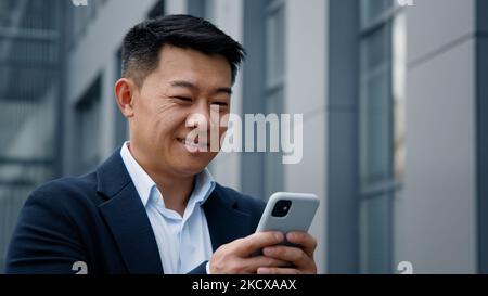 Close up smiling laughing Asian middle-aged Korean businessman man employer entrepreneur laugh looking at mobile phone chatting browsing smile Stock Photo