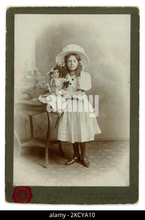 Edwardian era studio portrait cabinet card of pretty looking girl from a privileged, upper / middle class family wearing a large white hat, beautiful white dress (party ?)  with puffed sleeves, white gloves and tights and holding her china doll. Perhaps a birthday photograph. On reverse is written Clara R. J. Whant, aged 11 1909. From the studio of Edward Sharp Islington and Westminster Bridge London, U.K. Dated 1909. Stock Photo