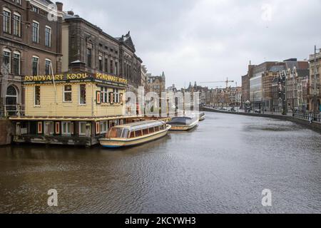 The empty canals of Amsterdam with the tourist sightseeing boats docked. Quiet, almost deserted from local people and tourists streets of Amsterdam during the lockdown in the Dutch capital city with stores and shops appearing with closed with the roller metal shutter down, cafes, bars and restaurants also closed with tables and chairs of the terraces locked. The Netherlands was the first European nation to declare full lockdown to fight the new Omicron variant that surges. After a sudden government order before Christmas, the country closed all the nonessential shops, cafes, restaurants, bars, Stock Photo