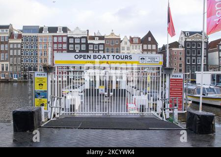 The empty canals of Amsterdam with the tourist sightseeing boats docked. Quiet, almost deserted from local people and tourists streets of Amsterdam during the lockdown in the Dutch capital city with stores and shops appearing with closed with the roller metal shutter down, cafes, bars and restaurants also closed with tables and chairs of the terraces locked. The Netherlands was the first European nation to declare full lockdown to fight the new Omicron variant that surges. After a sudden government order before Christmas, the country closed all the nonessential shops, cafes, restaurants, bars, Stock Photo