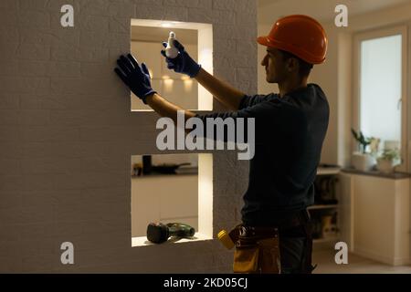 Handsome young builder in construction helmet is twisting the light bulb in. The man is looking up. Stock Photo