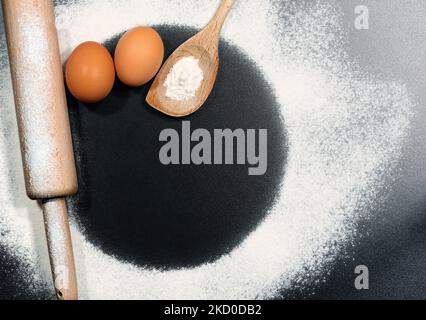 Baking ingredients on a black table with copy space, circle shape. Wooden rolling pin and spoon with white flour and two eggs. Stock Photo