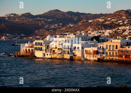 Panoramic view of Little Venice in Mykonos island during the magic hour of the sunset. Tourists enjoy a drink at the terrace or balcony of the whitewashed cafe bars or dinner at a restaurant just above the water at the little waterfront area under the famous windmills of the island. The Greek island of Myconos is a popular glamorous Mediterranean travel destination for holidays in the Cyclades, the Aegean Sea with the iconic whitewashed buildings, the sandy beaches and famous parties at the beach bars. The tourism and travel industry had a negative impact on the business and local economy sect Stock Photo