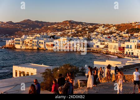 Panoramic view of Little Venice in Mykonos island during the magic hour of the sunset. Tourists enjoy a drink at the terrace or balcony of the whitewashed cafe bars or dinner at a restaurant just above the water at the little waterfront area under the famous windmills of the island. The Greek island of Myconos is a popular glamorous Mediterranean travel destination for holidays in the Cyclades, the Aegean Sea with the iconic whitewashed buildings, the sandy beaches and famous parties at the beach bars. The tourism and travel industry had a negative impact on the business and local economy sect Stock Photo