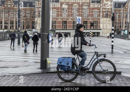 A woman as seen riding her bike with the Amsterdam central station in the background. Daily life during the lockdown at the fourth wave of the pandemic. Locals and a few tourists at the quiet streets of Amsterdam during the lockdown in the Dutch capital city with stores and shops appearing with closed with the roller metal shutter down, cafes, bars and restaurants also closed with tables and chairs of the terraces locked. The Netherlands was the first European nation to declare full lockdown to fight the new Omicron variant that surges. After a sudden government order before Christmas, the cou Stock Photo