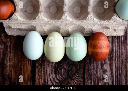 Horizontal Row of Blue, Green, and Brown Heritage Eggs With an Egg Carton: Multicolored eggs next to a paper egg carton on a wooden table Stock Photo