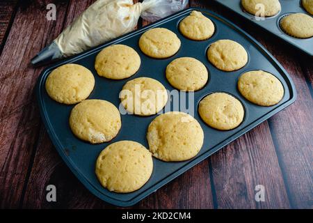Vanilla Cupcakes in a Muffin Pan with a Piping Bag of Buttercream: Vanilla cupcakes without frosting on a rustic wood table Stock Photo