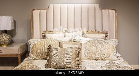 Elegant neutral satin bed cover and pillows against padded bed stead and beige wall with matching lamp Stock Photo