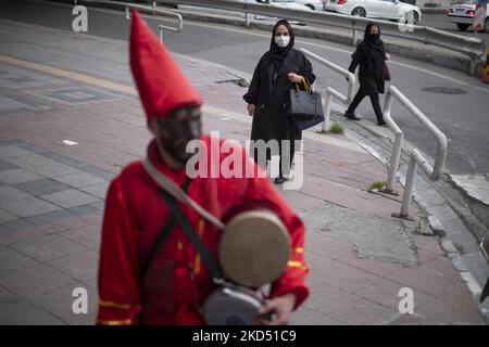 An Iranian woman wearing a protective face mask looks at a man who has painted his face in black and wore red dresses as Hajji Firuz (An Iranian New Year symbol), while standing on a street-side in northern Tehran on March 12, 2022. Hajji Firuz is a fictional character in Iranian folklore who appears in the streets by the beginning of Nowruz. His face is covered in soot, and he is clad in bright red clothes. He dances through the streets while singing and playing music. (Photo by Morteza Nikoubazl/NurPhoto) Stock Photo