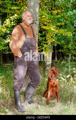 Casually in a country style with dungarees, sweater, rubber boots, an elderly man leans against a tree trunk. Stock Photo