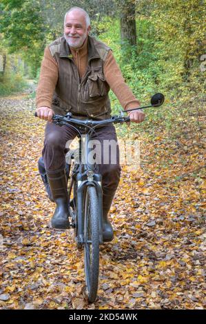 In a country style outfit, simple and relaxed, an active senior rides his electric bike through the colorful autumn forest. Stock Photo