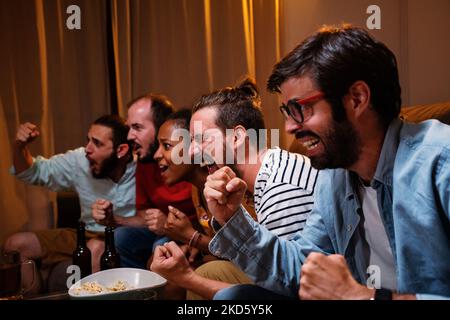 Group of five young friends watching sports on TV, cheer and celebrate. Happy diverse supporters fans sit on couch screaming excited with popcorn and Stock Photo