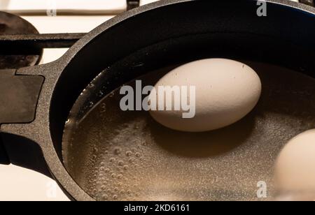 White chicken egg close-up with bubbles of boiling water in black pot on gas stove Stock Photo