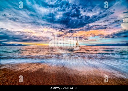 A Sailboat Sails Along The Ocean With A Colorful Cloudscape Overhead And Sun Rays Bursting Forth Stock Photo
