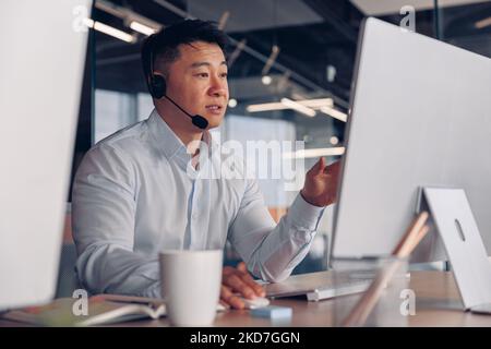 Focused asian businessman in headphones talking online with client sitting in office Stock Photo