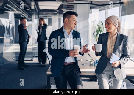 Two happy multiracial colleagues talking and drinking coffee during break time in office Stock Photo