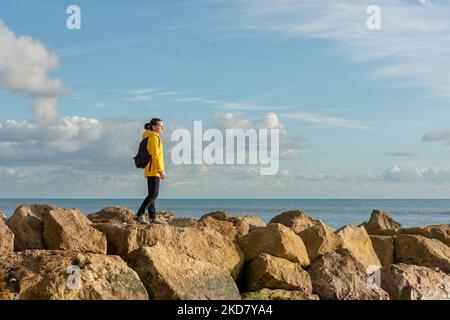 Woman wearing a yellow coat and backpack walking across rocks by the sea Stock Photo