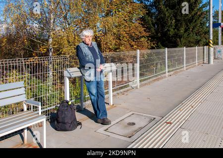 Old gray-haired bored tourist in blue jeans leaning on a wooden modern leaning bench or leaning stand or lean bar (no one name yet) waiting with backp Stock Photo