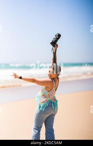 A vertical shot of a cheerful young woman with a professional camera at a beach on a bright sunny day Stock Photo