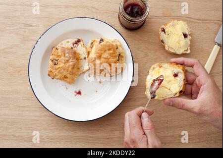 First person perspective view on pair of male hands applying jam on scones next to plate over table Stock Photo