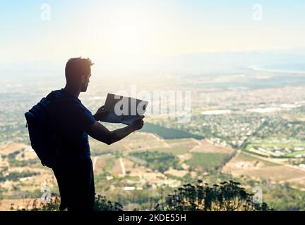 Keep going, keep exploring. Rear view shot of a young man reading a map while standing at the top of a mountain. Stock Photo