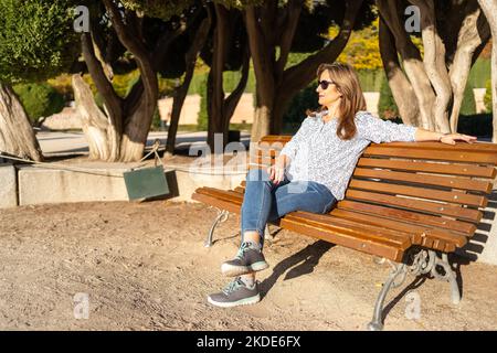 Senior woman sitting on a bench in a public park and enjoying the sun relaxedly. Stock Photo