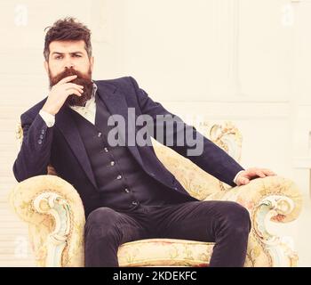 Macho attractive and elegant on serious face and thoughtful expression. Man with beard and mustache wearing classic suit, sits on old fashioned Stock Photo