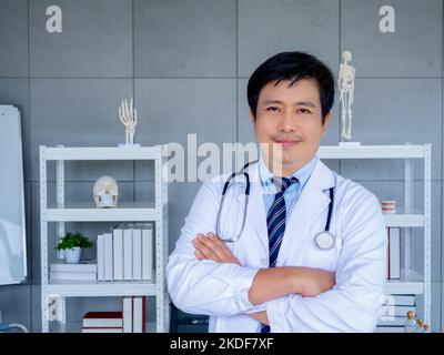 Smiling Asian man orthopedic doctor portrait in white coat standing with crossed arms near bookshelf and equipment in medical office. Confident adult Stock Photo