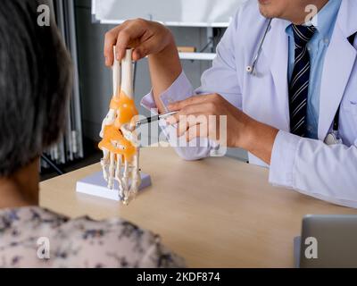 Close-up pen holding by orthopedic doctor man's hand in white coat pointing to foot skeleton, ankle joint anatomy model on desk to explain for patient Stock Photo