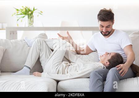 Woman lay head on boyfriend lap looking in each other eyes smile. Romantic husband and wife talking relaxing at home living room. Young affectionate m Stock Photo