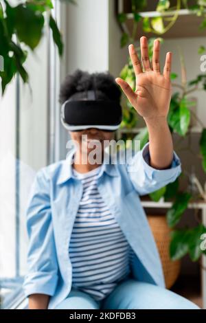 African woman in VR helmet interacting with objects in virtual world, enjoying immersive experience Stock Photo