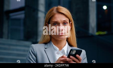 Serious focused businesswoman holding phone standing in city sad frustrated woman receiving email message reading bad news loses in online bet upset Stock Photo